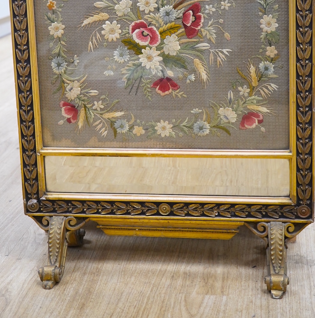 A late 19th century gilt framed silk and wool work embroidered fire screen, embroidered with a central sprig of poppies, wheat and wild flowers in a circular border of similar flowers, the frame with a decorative carved